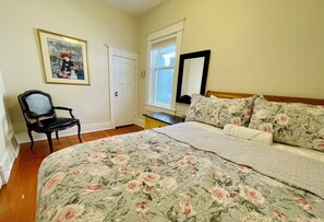Quarto com colcha florida, uma cadeira de madeira, mesa de cabeceira, espelho e porta.