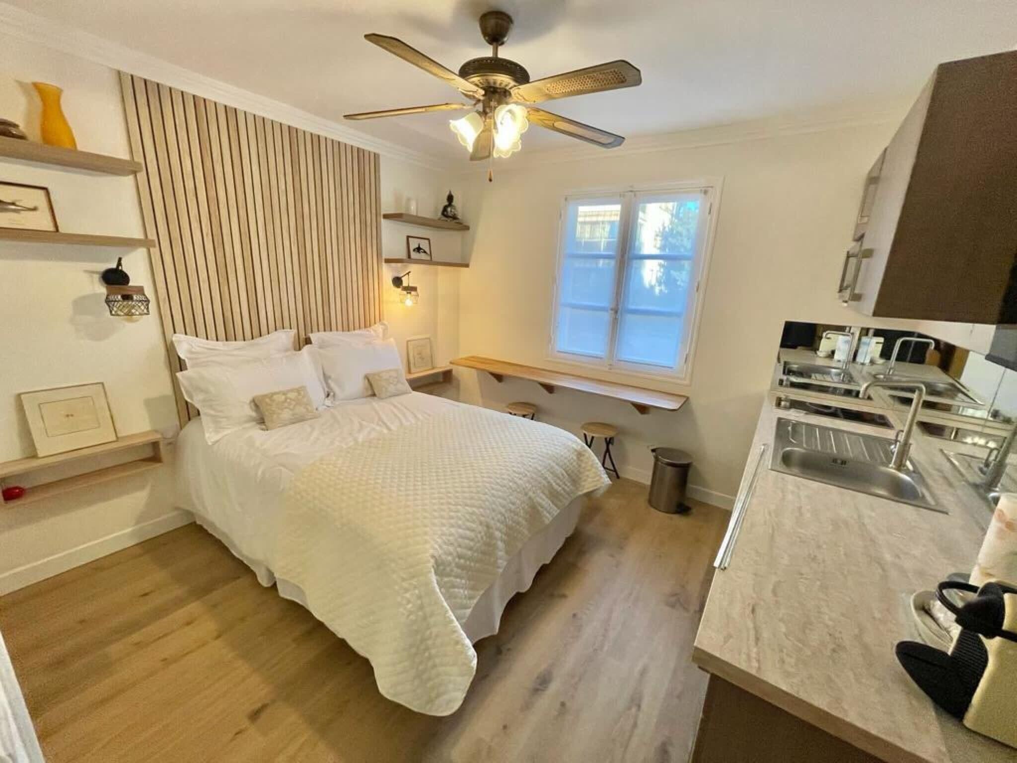A bedroom with a bed, bedside tables, a ceiling fan, a window, and a kitchenette with a sink.