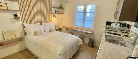 A bedroom with a bed, bedside tables, a ceiling fan, a window, and a kitchenette with a sink.