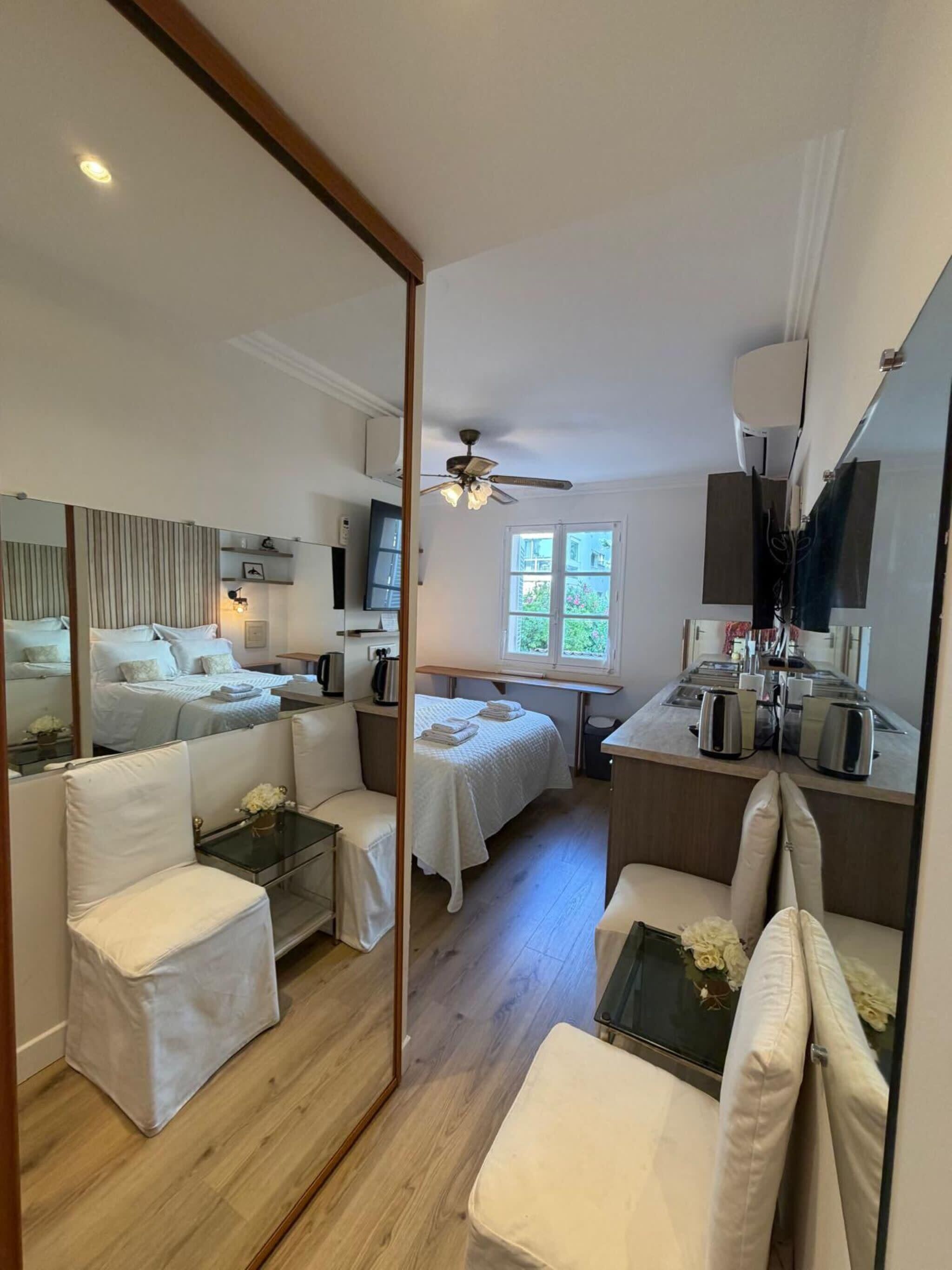 A hotel room with a bed, a sitting area with a chair, a small table, a mirror, a ceiling fan, and a window with a view of greenery.