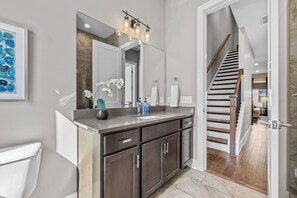 A modern bathroom with a large mirror, dark cabinetry, and a visible staircase leading to another level. - Lux Home Close to Broadway • Rooftop Views & FirePit! (Nashville)
