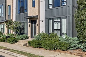 A modern multi-story residential building with a brick facade, large windows, and a well-maintained front yard with shrubs and a small tree. - Lux Home Close to Broadway • Rooftop Views & FirePit! (Nashville)