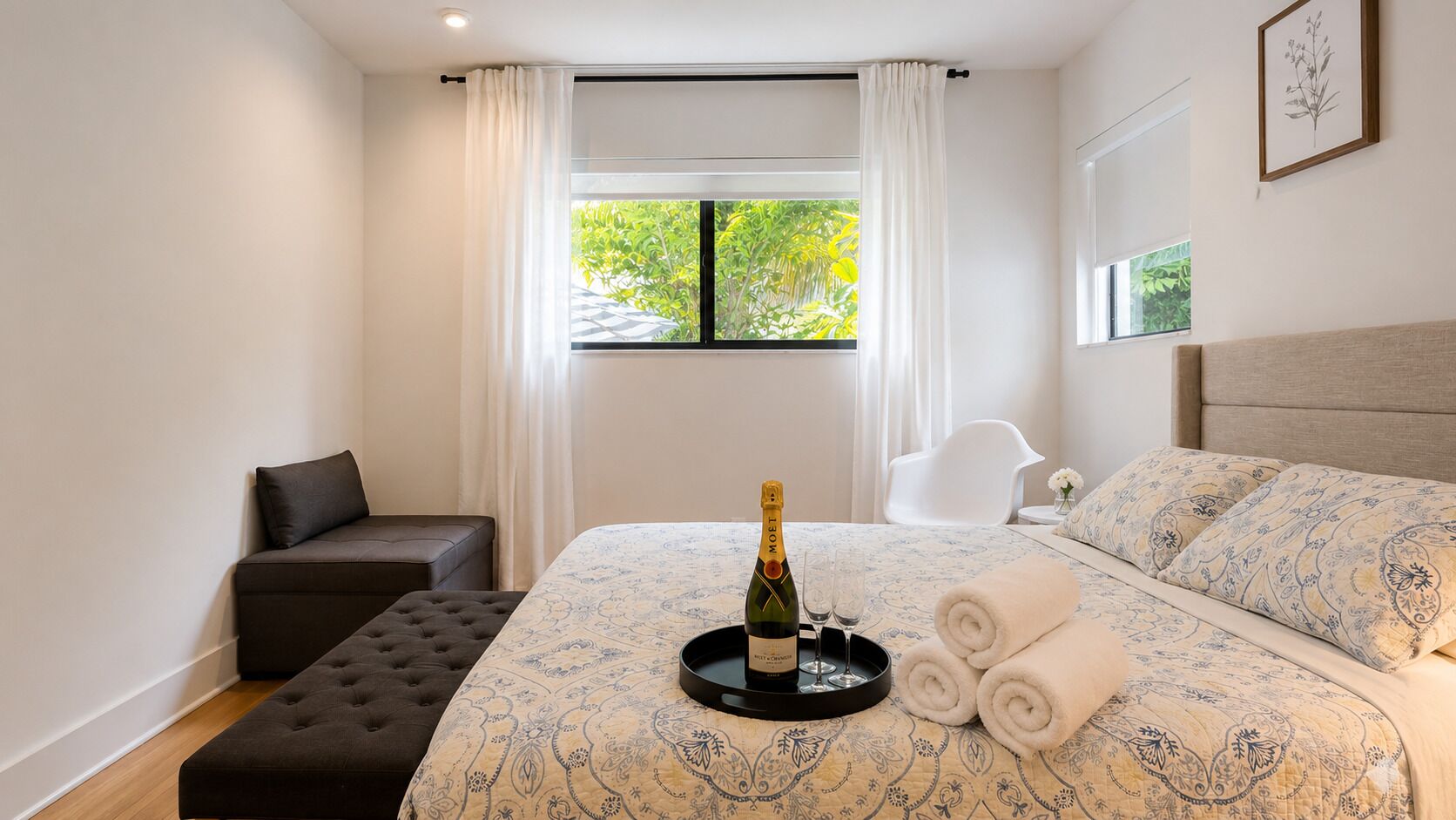 A hotel room with a bed, a small seating area, a window with curtains, and a bottle of champagne on a tray.