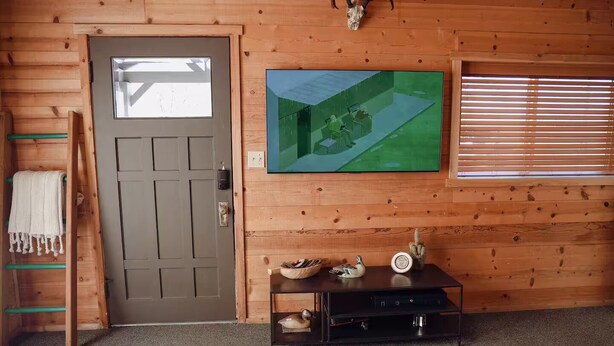 Ein Zimmer mit Holztür, einem wandmontierten Fernseher, einem kleinen Tisch mit Dekoration und einem Fenster mit Jalousien.