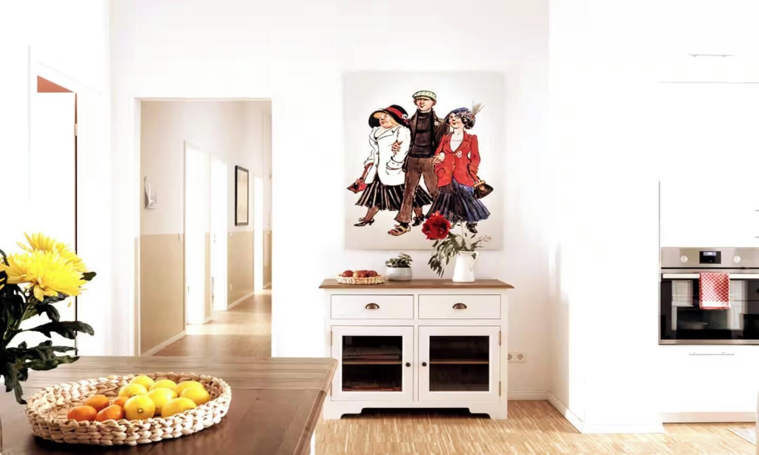 Eine moderne Küche mit weißen Schränken, einem Bild an der Wand und einer Obstschale auf dem Tisch.