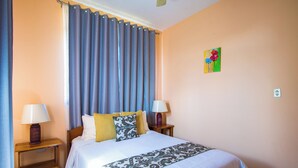 Quarto de hotel com cama, duas luminárias de cabeceira, janela com cortinas, um quadro na parede e uma tomada.