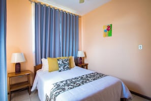 Ein Hotelzimmer mit einem Bett, zwei Nachttischlampen, einem Fenster mit Vorhängen, einem Wandbild und einer Steckdose.