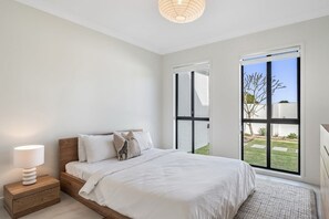Ein Schlafzimmer mit einem großen Bett, einem Nachttisch, einer Lampe und einem Fenster mit Blick auf einen Garten.