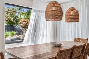 Ein Essbereich mit Holztisch und gewebten Stühlen, einer gewebten Pendelleuchte und Blick auf einen Garten durch transparente Vorhänge.