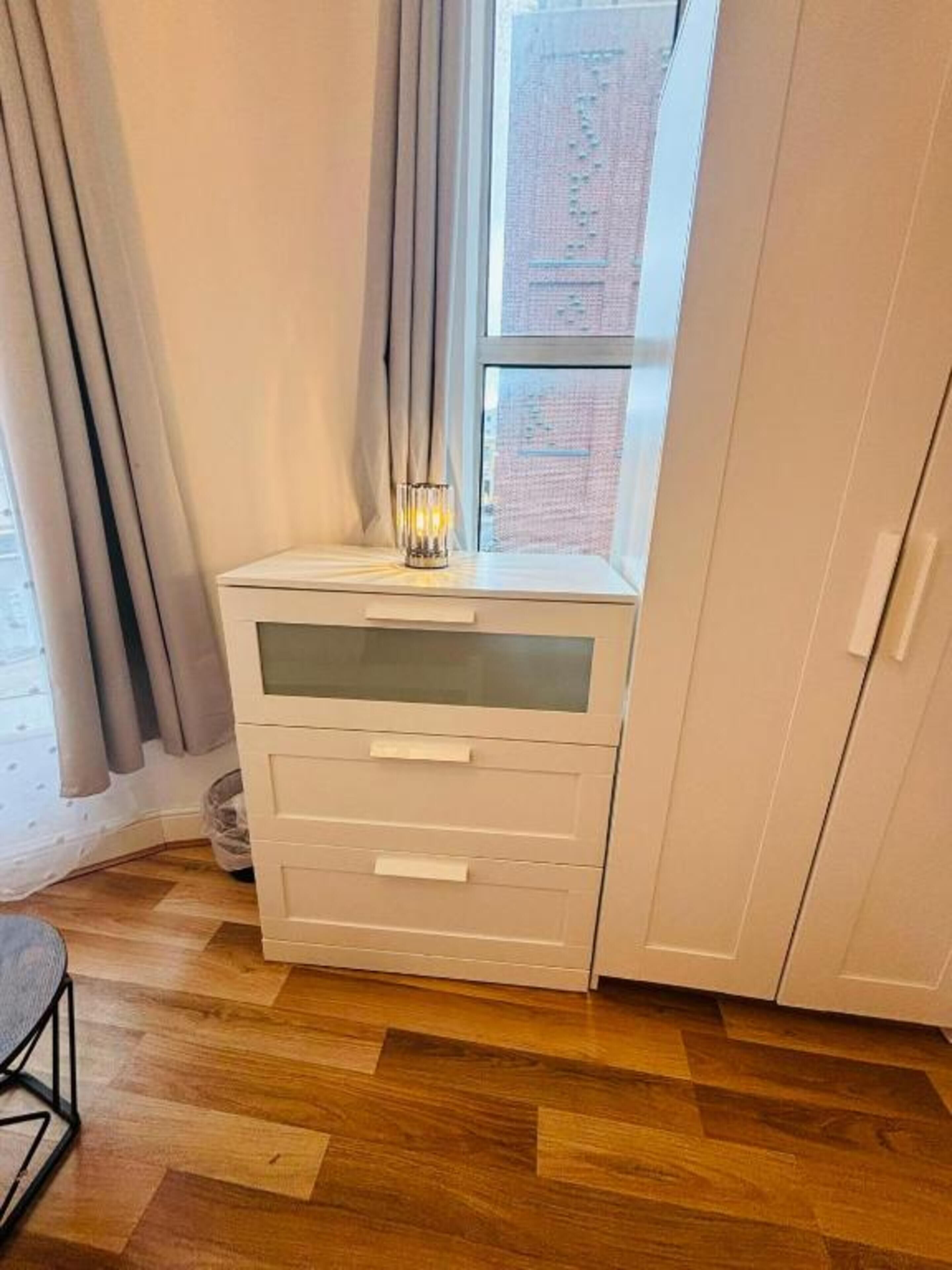 A corner bedroom with a wooden floor, a white bedside table with drawers, a window with curtains, and a view of a brick building.