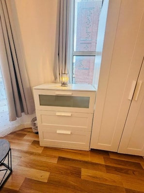 A corner bedroom with a wooden floor, a white bedside table with drawers, a window with curtains, and a view of a brick building.