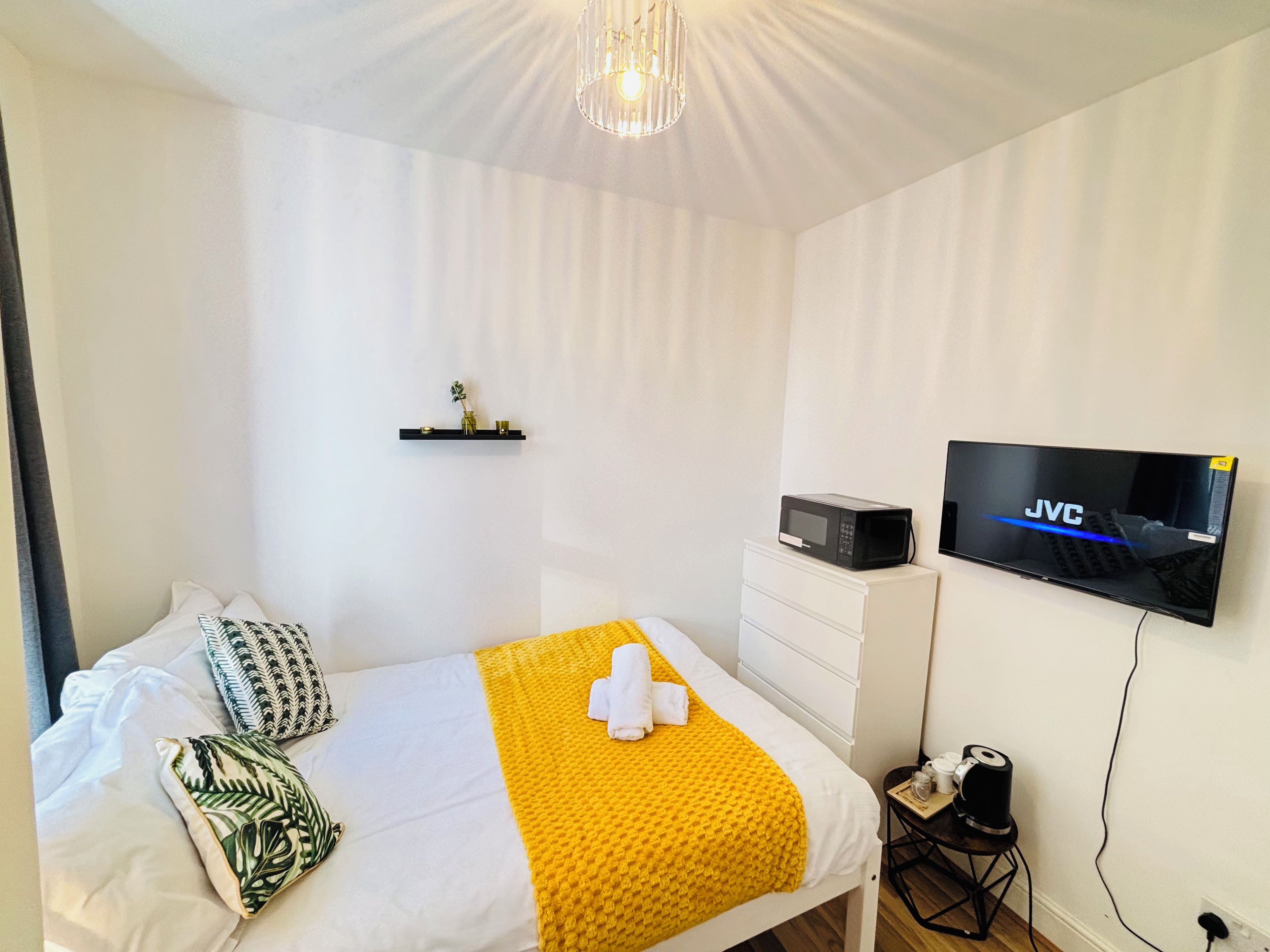 Hotellihuone, jossa on sänky, taulutelevisio, pieni pöytä, jossa on vedenkeitin, sekä hylly koriste-esineille.