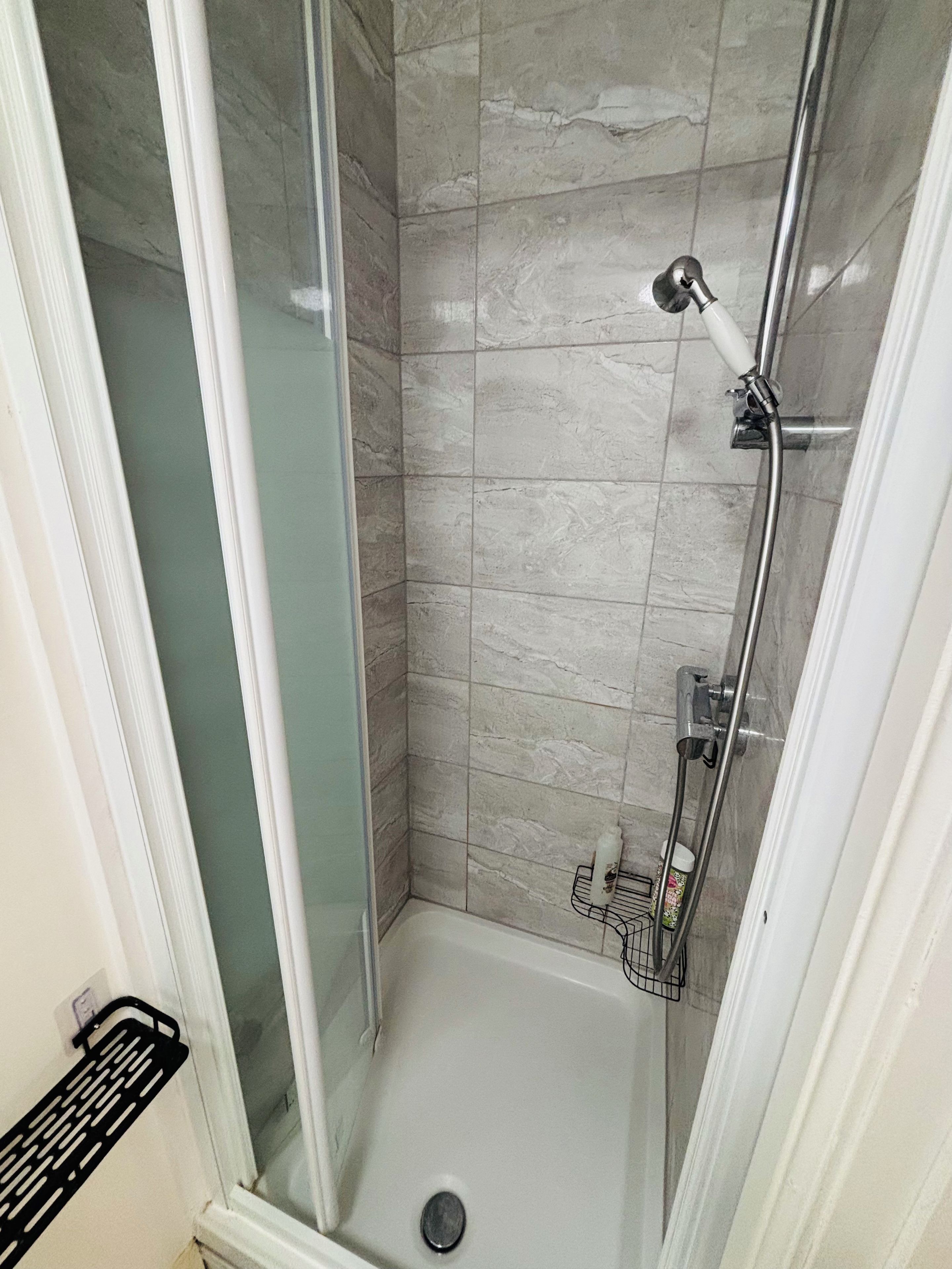 Een douche met een glazen deur, een handdouche en een metalen rek met een fles.