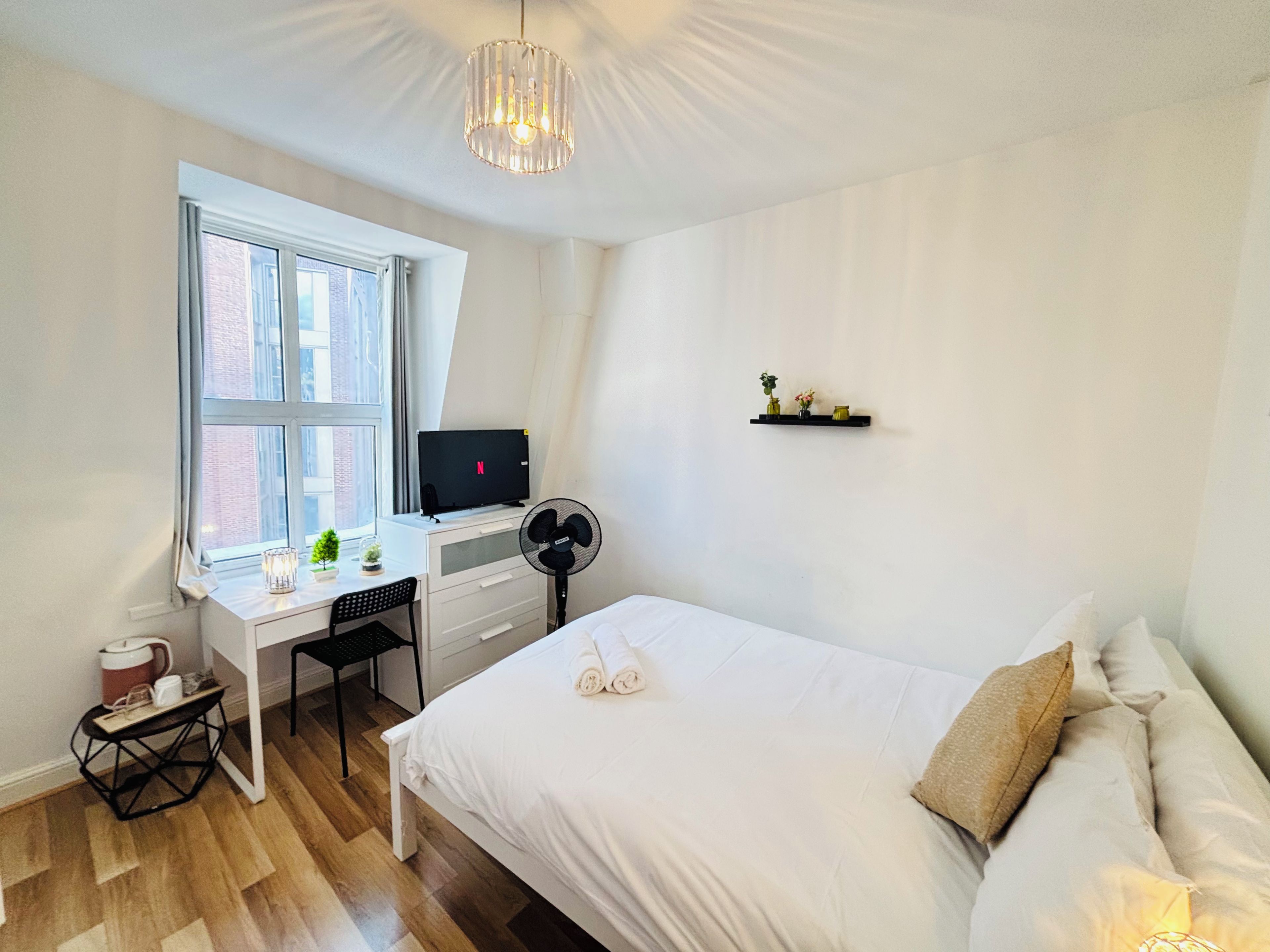 A bedroom with a bed, a desk with a computer, a fan, and a window.