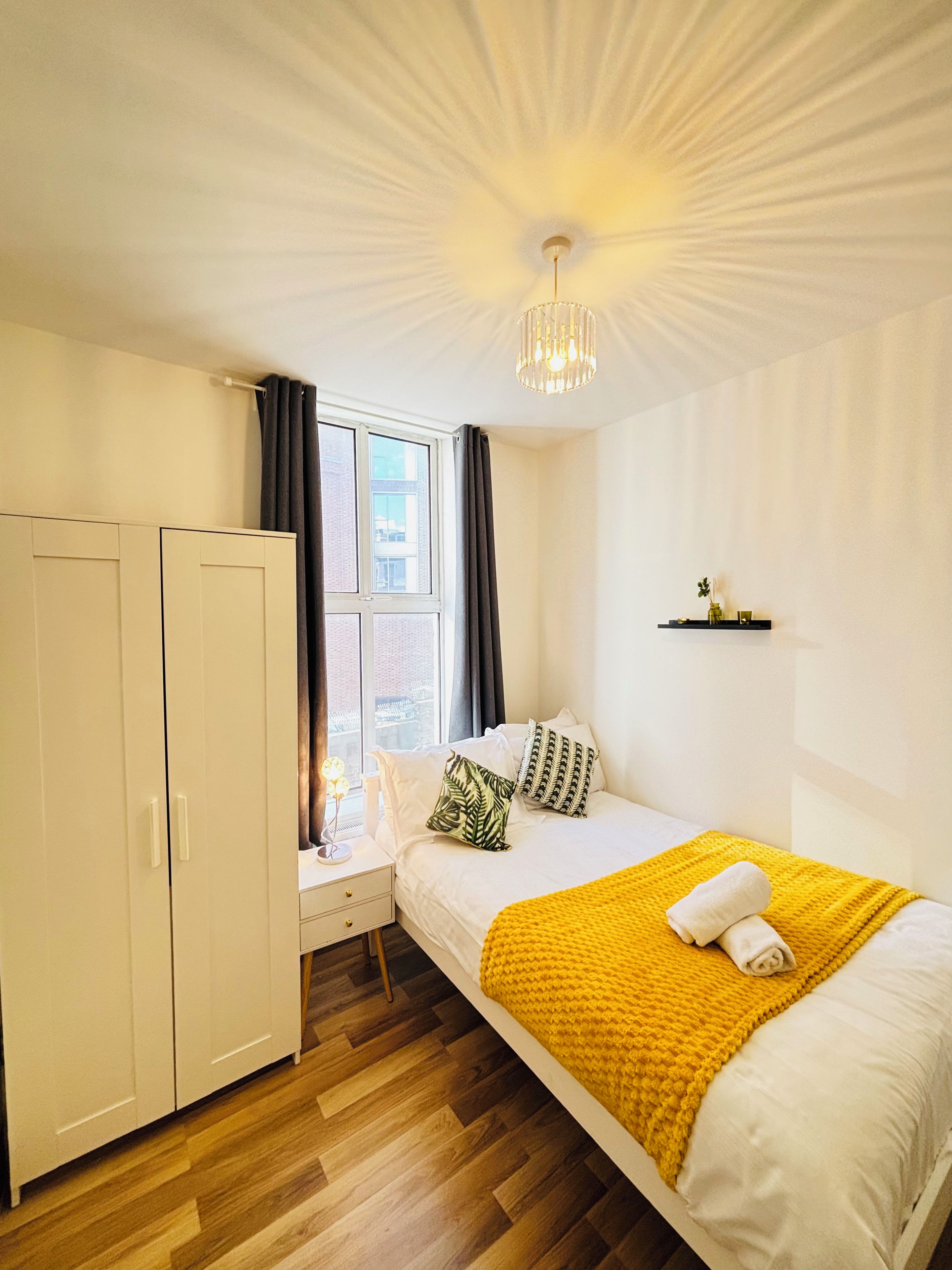 Un dormitorio con cama, armario, mesita de noche, ventana con cortinas y lámpara en el techo.