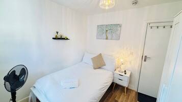 Een slaapkamer met een bed, nachtkastje, lamp, ventilator, kunstwerk en een deur.