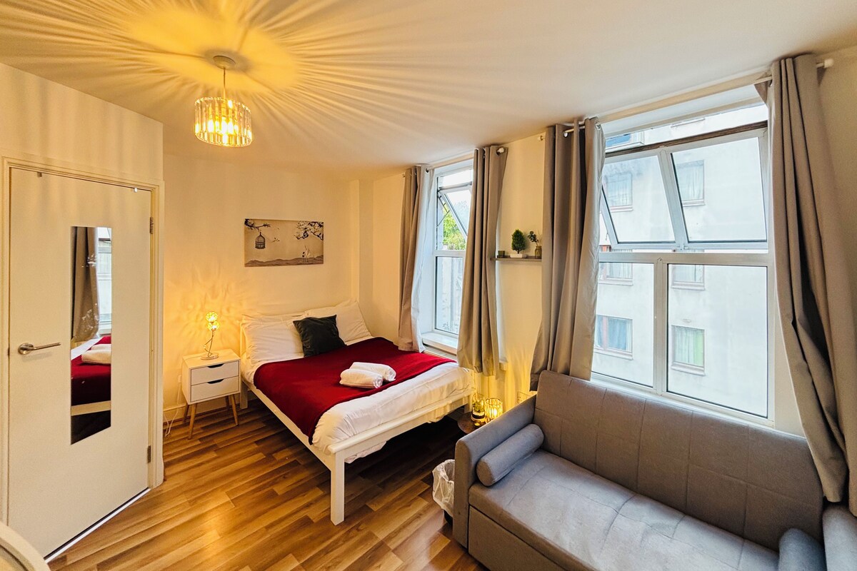 Habitación de hotel con cama, sofá, mesita de noche, lámpara, ventana con cortinas y un cuadro en la pared.