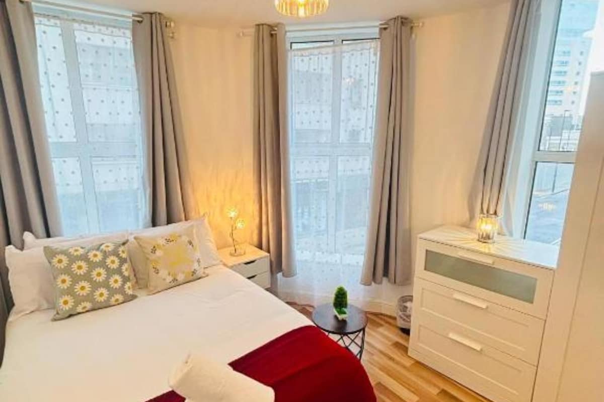 Hotellihuone, jossa on sänky, yöpöytä, lamppu ja suuret ikkunat, joista avautuu näkymä kaupunkiin.