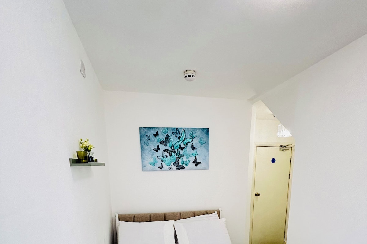 Una habitación de hotel con una cama, un cuadro en la pared, una puerta y un pequeño estante con un jarrón.