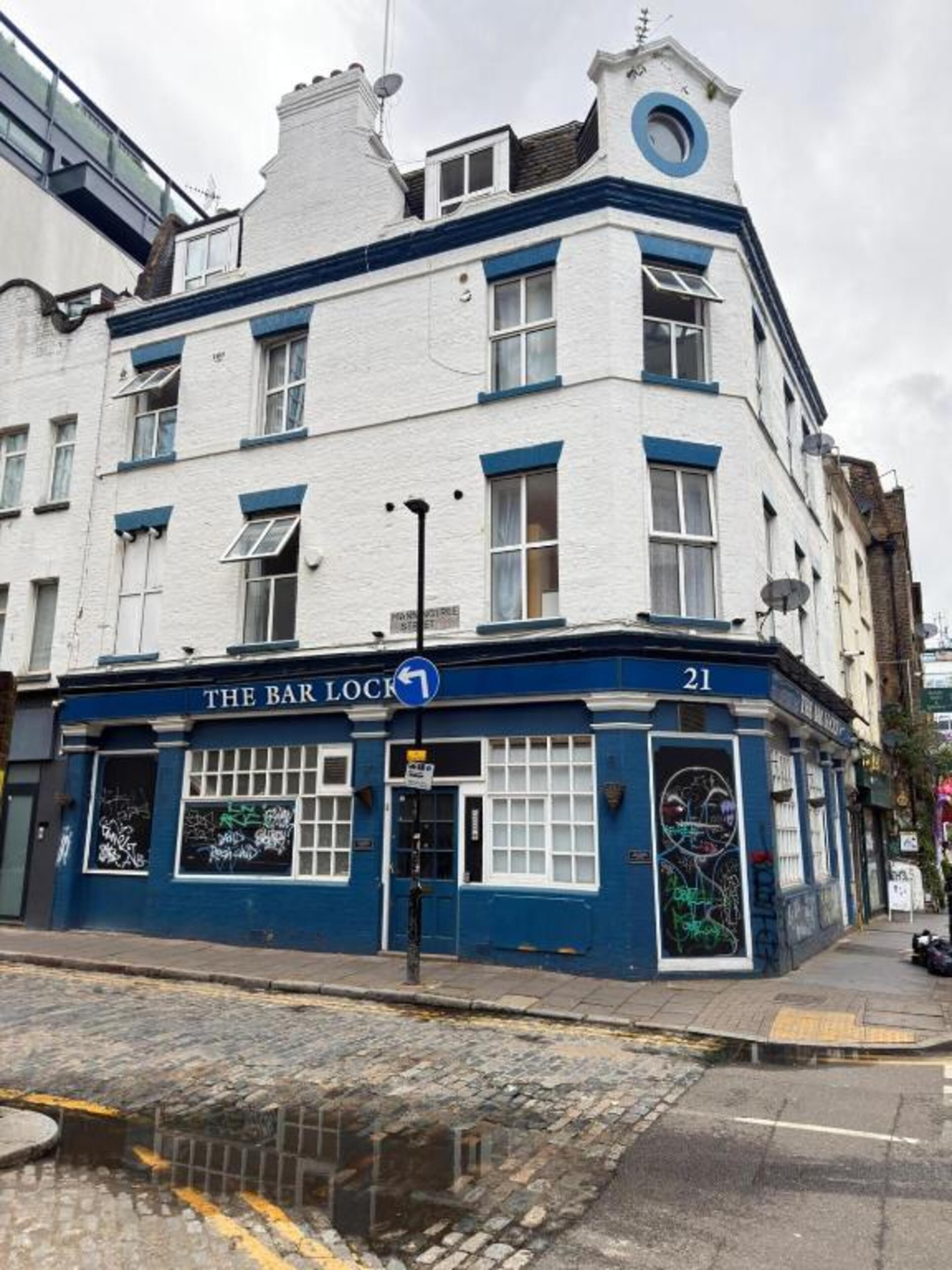 A building with a blue and white facade, a sign reading 'The Bar Lock,' and a number '21' on the corner.