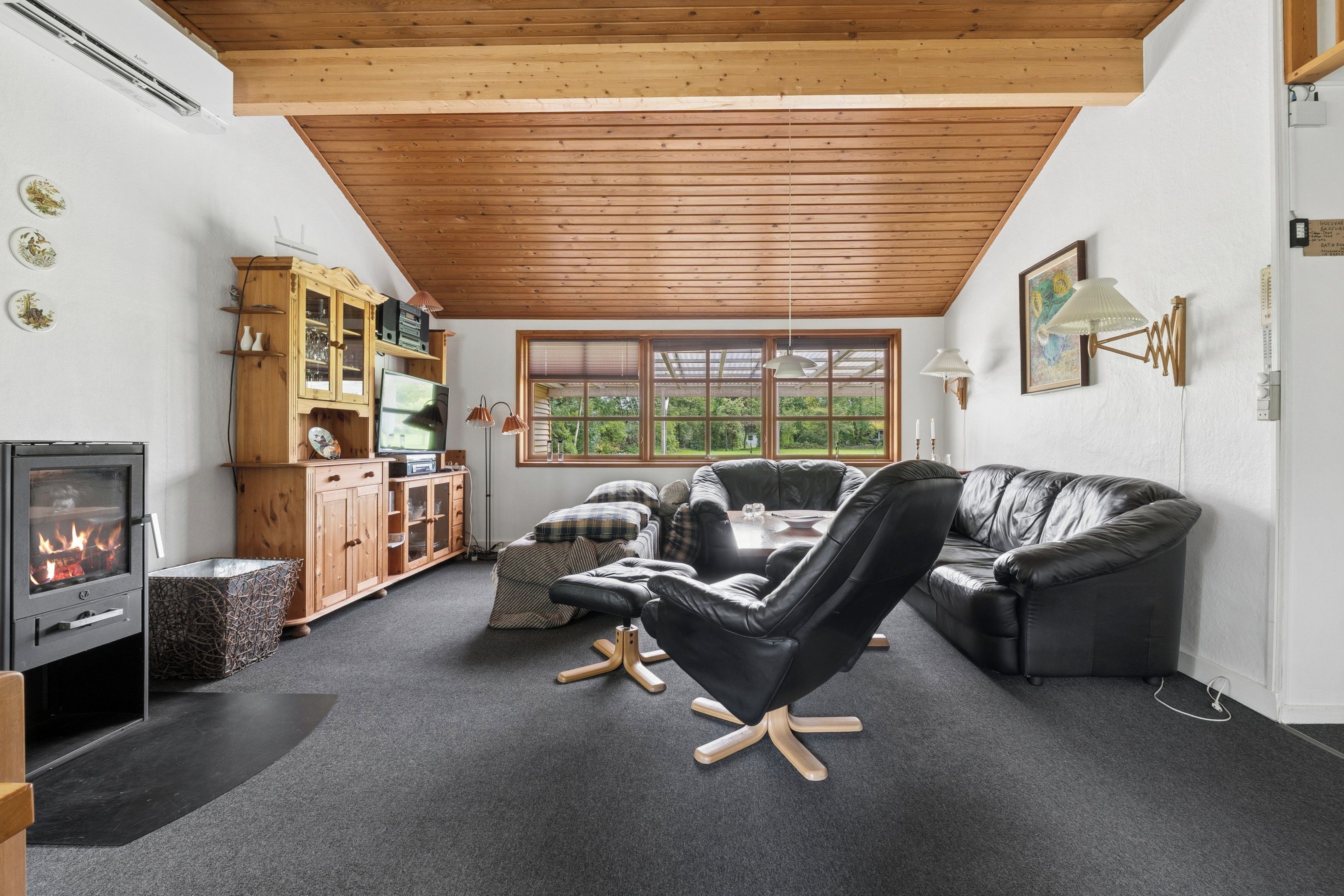 A cozy living room with a fireplace, a wooden cabinet, a black leather sofa, and a large window providing a view of greenery.