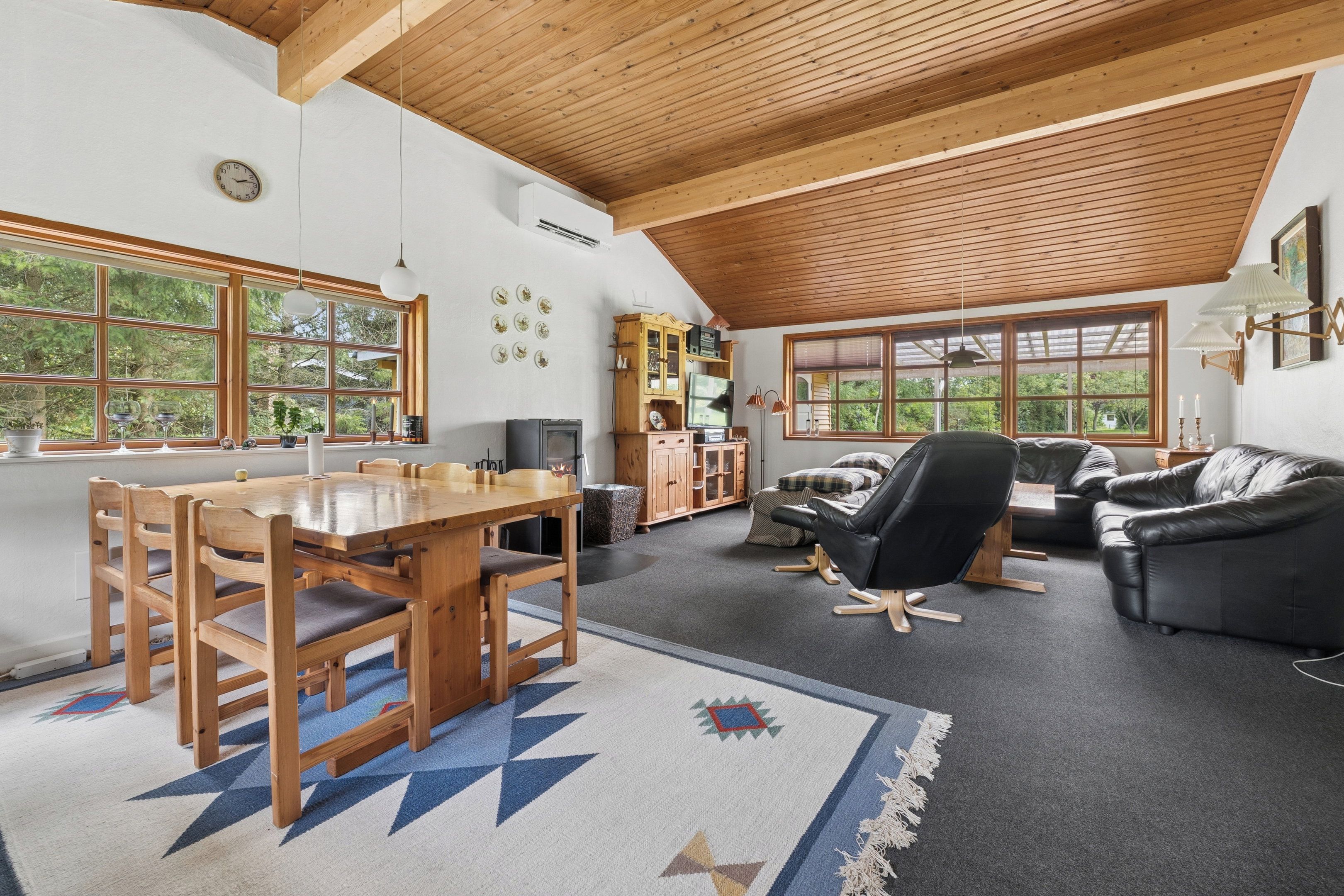 Amplia sala de estar con techo de madera, comedor con mesa y sillas de madera, sofá de cuero negro y un ventanal con vistas al exterior.