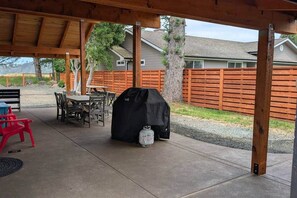 Ein überdachter Patio-Bereich mit Grill, Gartenmöbeln und einem Holzzaun im Hintergrund.
