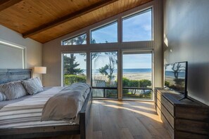 Ein Schlafzimmer mit großem Fenster mit Blick auf einen Strand, einem Bett mit gestreifter Bettwäsche und einem Fernseher auf einer Holzkommode.