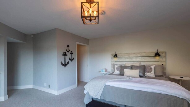 A bedroom with a bed, a nightstand, a wall-mounted light fixture, and a door leading to another room. - Devon West (Newport)