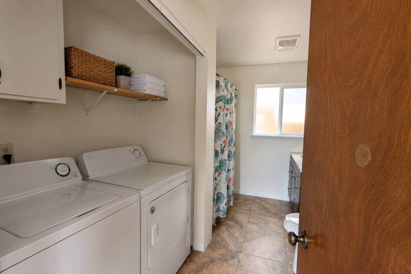 Un espace buanderie comprenant un lave-linge et un sèche-linge, une étagère avec des serviettes, ainsi qu’une salle de bain avec des toilettes et un rideau de douche.