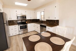 Una cocina moderna con armarios blancos, un refrigerador de acero inoxidable, un microondas, un horno y un lavavajillas. Hay una mesa de comedor de madera con cuatro individuales tejidos.
