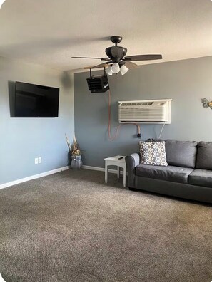 A living room with a gray couch, a small white table, a ceiling fan, a mounted TV, and an air conditioning unit. - Cozy 1-bedroom house in wonderful Doniphan with AC (Doniphan)