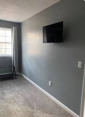 Ein Zimmer mit grauer Wand, einem an der Wand befestigten Flachbildfernseher, einem Fenster mit Vorhängen und einem Teppichboden.