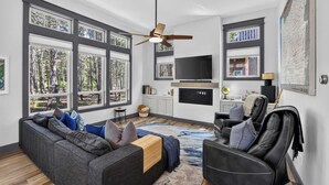 Un salon moderne avec un canapé gris, un fauteuil en cuir noir et une grande fenêtre donnant sur la forêt.