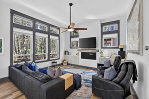 A modern living room with a grey sofa, black leather chair, and a large window with a view of trees. - Dory Coastal Paradise - walk to Cape Kiwanda (Pacific City)