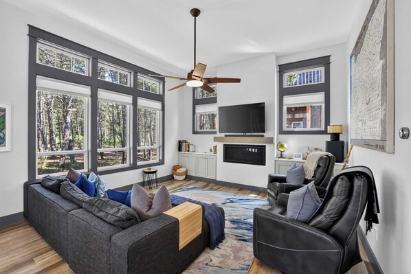 Ein modernes Wohnzimmer mit grauem Sofa, schwarzem Ledersessel und großem Fenster mit Blick auf Bäume.