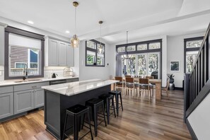 A modern kitchen with a central island, bar stools, and a dining area with a view of the outdoors. - Dory Coastal Paradise - walk to Cape Kiwanda (Pacific City)