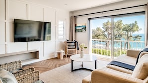 A modern living room with a flat-screen TV, wicker furniture, a round coffee table, and a view of the sea. - Camps Bay 2 Bed With Sea Views Shoreline Views (Cape Town)