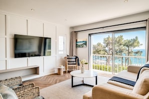 Ein modernes Wohnzimmer mit Flachbildfernseher, Korbmöbeln, einem runden Couchtisch und Blick auf das Meer.