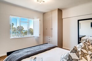 Ein Schlafzimmer mit großem Fenster, einem Bett mit gemusterten Betttextilien, einem Kleiderschrank und einem Spiegel.