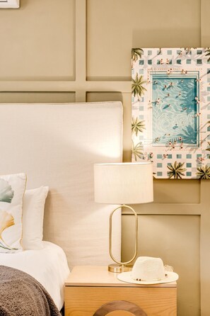 Una habitación de hotel con una cama, una mesita de noche con una lámpara, un sombrero y un cuadro con una vista de una piscina.