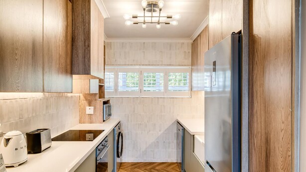 A modern kitchen with wooden cabinetry, a stainless steel refrigerator, a built-in oven, and a window providing natural light. - Camps Bay 2 Bed With Sea Views Shoreline Views (Cape Town)
