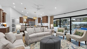 Ein modernes Wohnzimmer mit Sofa, Hocker und Sesseln, einer Küchenzeile sowie einem Balkon mit Blick auf einen Pool.