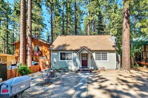 A single-story house with a front porch, surrounded by tall pine trees.