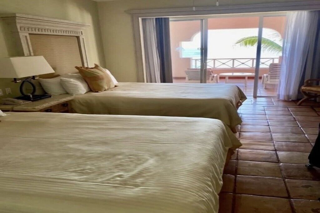 A hotel room with two beds, a nightstand, a lamp, and a view of the outdoor area through a sliding door.