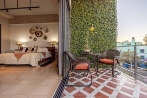 Een slaapkamer met een bed, nachtkastjes en decoratieve wandkunst. Een balkon met een groene wand en uitzicht op de stad is zichtbaar.