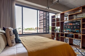 Ein modernes Hotelzimmer mit großem Fenster, einem Bett, einem Bücherregal und einem Flachbildfernseher.