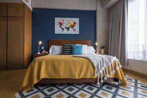 Ein Schlafzimmer mit Bett, Nachttisch, Lampe, Wandbild und einem Fenster mit Vorhängen.
