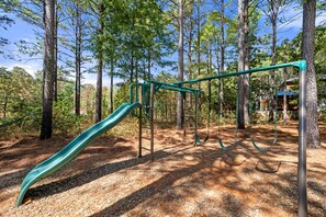 Ein Spielplatz mit Rutsche, Schaukeln und einer Kletterkonstruktion in einem Waldgebiet.