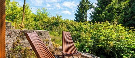 Two wooden lounge chairs in a garden with dense greenery and a clear sky.